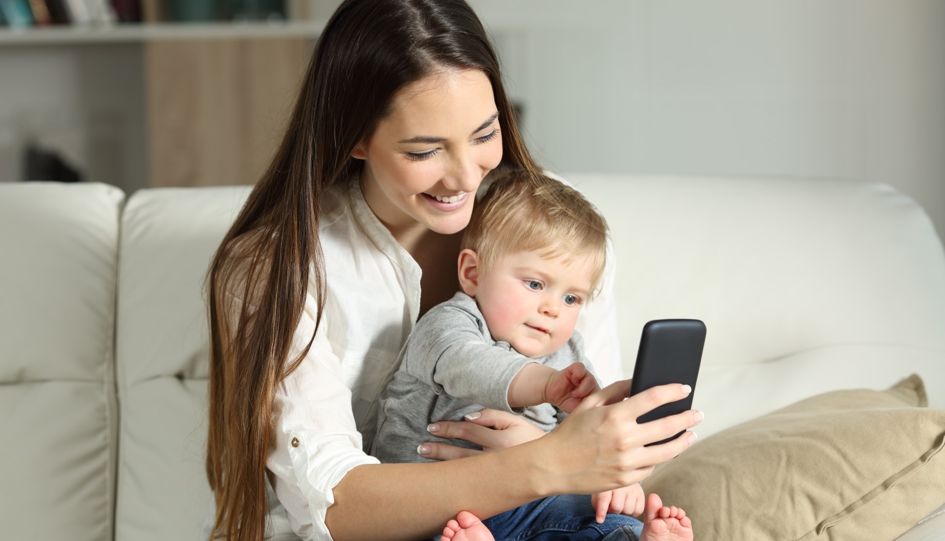 Parent holding toddler and looking at their phone with a smile whilst the child points to the phone